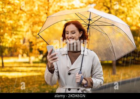 Lächelndes, hübsches kaukasisches, tausendjähriges Weibchen mit roten Haaren in Regenmantel und Regenschirm genießt den Regen und plaudert auf dem Smartphone Stockfoto