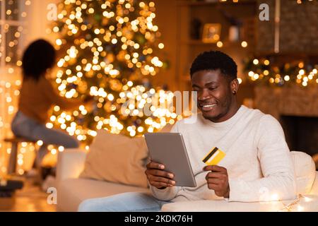 Froher Millennial schwarzer Mann mit Tablet und Kreditkarte Genießen Sie Online-Shopping, Frau schmückt Weihnachtsbaum Stockfoto