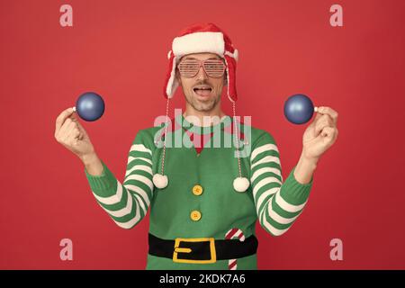 Staunender Mann im Elf Kostüm und weihnachtsmann Hut. Kerl in Party-Brille halten Weihnachten Dekoration Kugeln Stockfoto