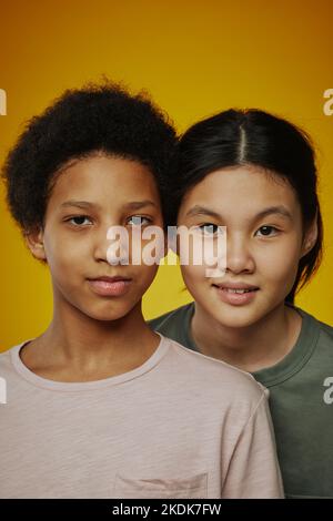 Portrait von zwei heranwachsenden freundlichen Mädchen in T-Shirts, die vor der Kamera auf hellgelbem Hintergrund stehen und auf Sie blicken Stockfoto