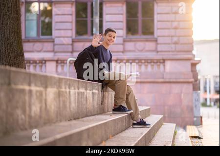 Fröhlicher Teenager saß auf der Treppe und begrüßte jemanden draußen Stockfoto