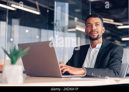 Schöner afrikanischer Geschäftsmann, der in einem modernen Büro sitzt und mit einem Laptop arbeitet und wegschaut Stockfoto