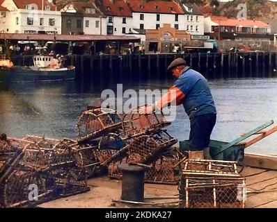 Eine alte Momentaufnahme eines Fischers aus Whitby, Yorkshire, der traditionelle handgemachte Krabben-/Hummertöpfe stapelt und in seiner selbstgemachten Schubkarre transportiert wird. Der Pier auf der Westseite des Flusses Esk, mit seinem Fischmarkt Gebäude kann mit einem lokalen Fischerboot in der Nähe vor Anker gesehen werden. Stockfoto