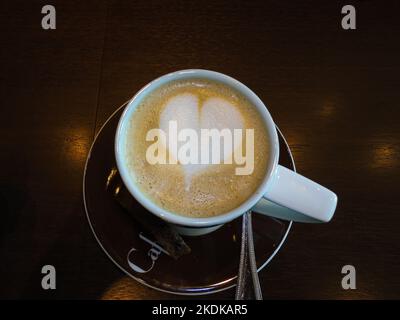 Nahaufnahme einer weißen Kaffeetasse mit herzförmiger Latte-Kunst auf einer Untertasse mit Utensil, die auf einem Holztisch ruht. Stockfoto
