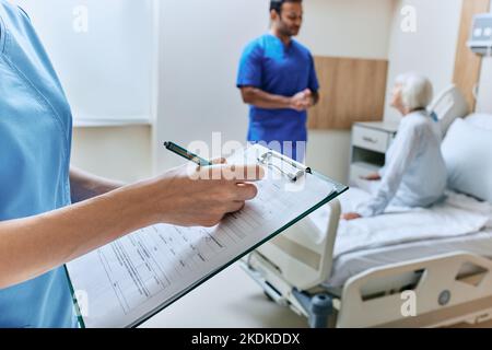 Die Krankenschwester macht Notizen in der Krankenakte im Krankenhauszimmer mit älteren Patienten und Arzt während der Beratung am Krankenbett. Medizinische Versorgung des Patienten Stockfoto