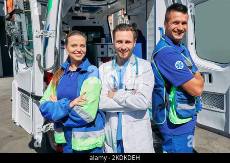 Rettungsteam. Selbstbewusstes Sanitäterteam und Krankenhausarzt mit gekreuzten Armen, die in der Nähe des Krankenwagens stehen Stockfoto