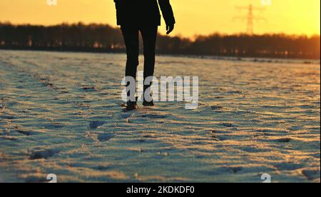 Schwarz gekleidete Person, die bei Sonnenuntergang durch ein schneebedecktes Feld geht Stockfoto