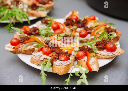 Tapas auf einem Teller. Brotscheiben mit Thunfisch-Creme, Kirschtomatenscheiben, mit gebratenem Speck bestreut und mit frischem arug garniert Stockfoto