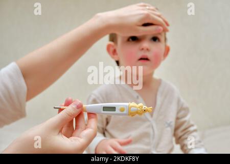 Eine Mutter misst die Temperatur eines kranken Kleinkindes mit einem Thermometer. Mama überprüft die hohe Temperatur eines kranken Kindes. Kind im Alter von einem Jahr Stockfoto