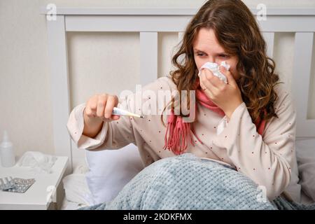 Eine kranke Frau bläst ihre Nase mit einem Thermometer in der Hand, Fieber und hoher Temperatur in eine Serviette. Erwachsene kranke Frau mit einem roten Schal auf einem ho sitzen Stockfoto