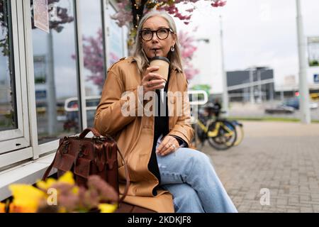 Ältere Frau, die Kaffee trinkt, sitzt auf einer Bank in der Nähe des Ladens Stockfoto