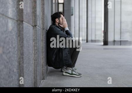 Ein Mann in einem depressiven Zustand sitzt zurückgelehnt an die Wand und bedeckt seine Hände mit seinen Handflächen Stockfoto
