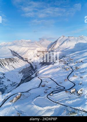 Luftaufnahme der verschneiten Bergkette bei Wintersonnenaufgang im Skigebiet. Drohne über Bergtal und Dorf mit kurviger Straße bei Sonnenuntergang. Gipfel des Kaukasus Stockfoto