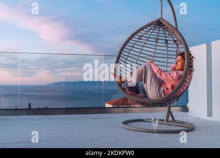 Schöne Frau mittleren Alters, barfuß, die in einem bequemen Hängesessel auf der offenen Terrasse liegt und die Sonnenstrahlen des Sonnenuntergangs genießt. Moderner Lebensstil zu Hause Stockfoto