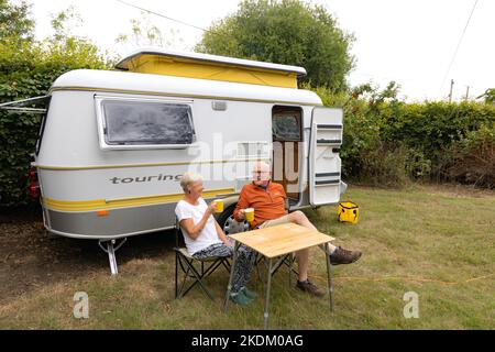 Karawanenurlaub in Großbritannien; Ein reifes Senioren-Paar im Ruhestand sitzt vor ihrem gelben ERIBA-Wohnwagen und genießt einen Drink. Concept Retirement UK Stockfoto