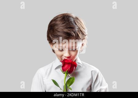 Bezaubernder eleganter weißer Junge mit einer roten Rose als Geschenk für seine Mutter oder Freundin. Muttertag oder Valentinstag Konzept Stockfoto