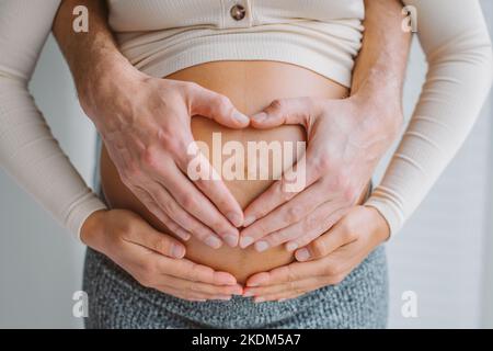 Liebevolle Eltern Herz Form Hände auf schwanger Frau Bauch. Erwartet Paar in der Liebe Schwangerschaft Nahaufnahme Foto des Bauches. Stockfoto
