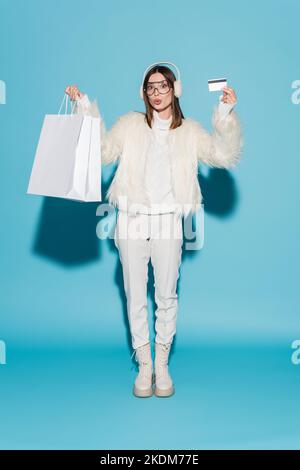 In voller Länge überrascht Frau in Ohrenschützer und weißen Kunstpelzjacke mit Kreditkarte und Einkaufstaschen auf blau Stockfoto