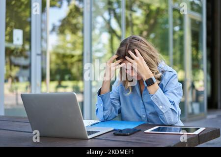 Porträt einer jungen arbeitenden Frau, die in der Nähe des Laptops sitzt und sich frustriert und enttäuscht fühlt. Geschäftsfrau sieht beunruhigt aus, während sie an einem Projekt arbeitet, sitzt Stockfoto