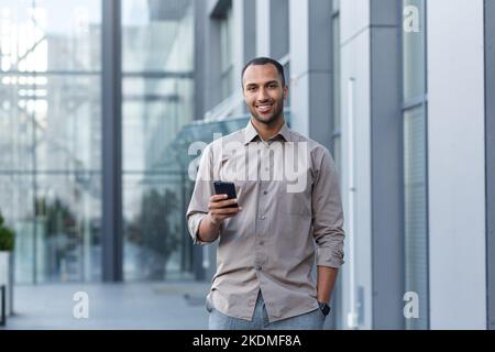 Porträt eines afroamerikanischen Geschäftsmannes, eines Mannes, der das Telefon lächelt und die Kamera anschaut, eines Büroarbeiters im Hemd vor dem modernen Bürogebäude in der Mittagspause. Stockfoto