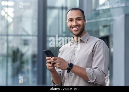 Porträt eines afroamerikanischen Geschäftsmannes, eines Mannes, der das Telefon lächelt und die Kamera anschaut, eines Büroarbeiters im Hemd vor dem modernen Bürogebäude in der Mittagspause. Stockfoto