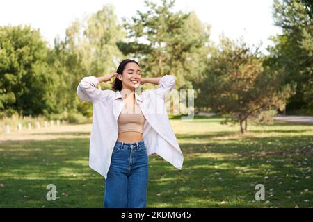 Sorgloses asiatisches Mädchen, das im Park lacht und tanzt, genießt den sonnigen Sommertag, hebt die Hände hoch und atmet frische Luft Stockfoto