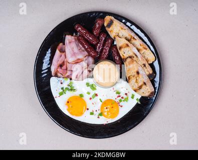 Englisches Frühstück. Speck, Spiegeleier, Würstchen, Sauce, geröstetes Brot. Stockfoto