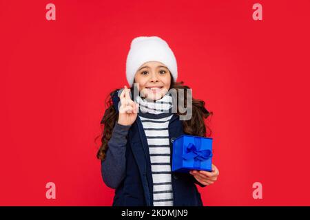 Glückliches Kind in Kugeljacke und Hut halten Box. Kind mit Geschenk. Teen Mädchen auf rotem Hintergrund. Winterferien. Crossing Finger, um einen Wunsch zu machen. Stockfoto