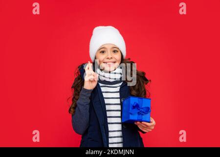 Glückliches Kind in Kugeljacke und Hut halten Box. Kind mit Geschenk. Teen Mädchen auf rotem Hintergrund. Winterferien. Crossing Finger, um einen Wunsch zu machen. Stockfoto