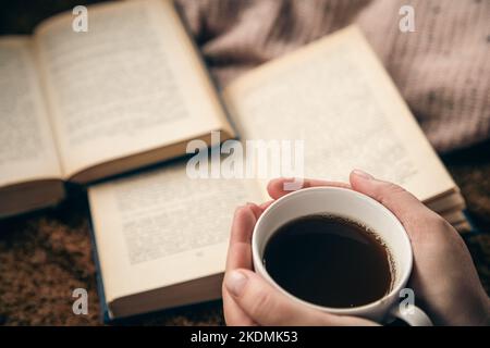 Nahaufnahme, eine Tasse Tee in weiblichen Händen auf einem verschwommenen Buchhintergrund. Stockfoto