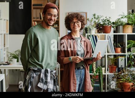 Zwei glückliche junge interkulturelle Programmierer in Casualwear, die Sie lächelnd anblicken, während einer von ihnen während der Teamarbeit einen Laptop in der Hand hält Stockfoto
