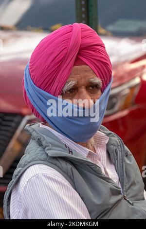 Posierte Porträt eines älteren Sikh-Mannes in einem rosa Turban und Teilmaske. An der 74. Street in Jackson Heights, Queens, New York City. Stockfoto