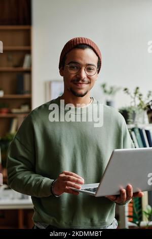 Junger, selbstbewusster IT-Techniker in der Freizeit schaut im Büro auf die Kamera, während er einen Laptop verwendet und mit Online- oder kodierten Daten arbeitet Stockfoto
