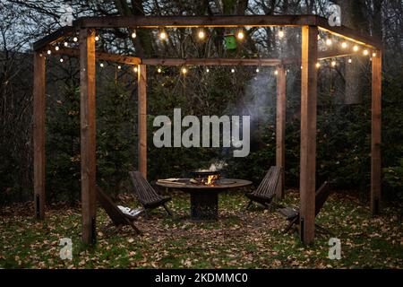 Hölzerne Gartenpergola mit Grill in der Dämmerung, Dekorlampe und Stühle im Herbst. Stockfoto