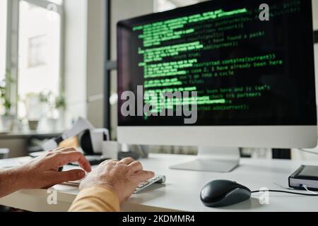 Konzentrieren Sie sich auf die Hände eines reifen Programmierers, der auf der Tastatur tippt, während Sie vor dem Computermonitor mit kodierten Daten auf dem Bildschirm sitzen Stockfoto