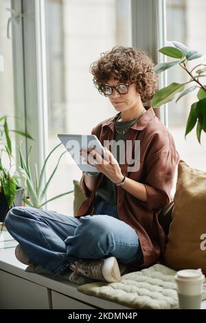 Junge, seriöse Geschäftsfrau in Casualwear, die im Büro auf einer Fensterbank zwischen Grünhauspflanzen sitzt und ein digitales Tablet verwendet Stockfoto