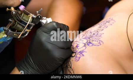 Nahaufnahme, in einem Tattoo-Salon , ein Spezialist macht ein Tattoo auf dem Rücken der Frau, schwarze Farbe floralen Ornament. Ein Mann arbeitet in speziellen schwarzen Handschuhen, auf spezielle Ausrüstung. Hochwertige Fotos Stockfoto