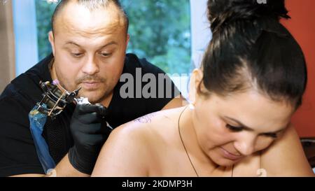 In einem Tattoo-Salon tätowiert ein Spezialist auf dem Rücken der Frau, einem Blumenschmuck. Ein Mann arbeitet mit speziellen Handschuhen, auf Spezialausrüstung. Zeichnen wird mit schwarzer Farbe gemacht. Hochwertige Fotos Stockfoto