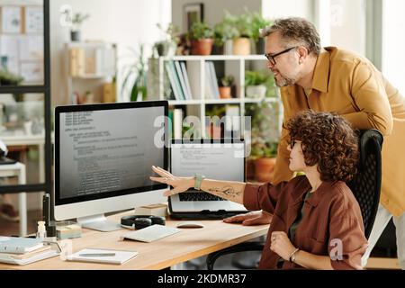 Junger selbstbewusster Programmierer, der auf Daten auf dem Computerbildschirm zeigt, während er mit Kollegen Brainstorming und Möglichkeiten der Dekodierung diskutiert Stockfoto