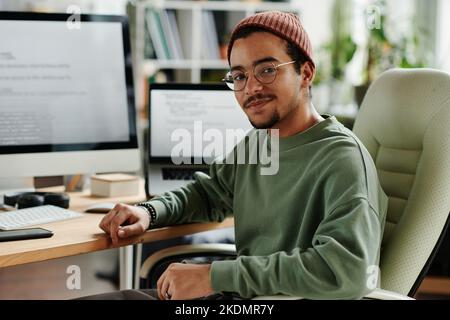 Junge, selbstbewusste IT-Ingenieure oder Softwareentwickler in Casualwear sitzen im Sessel am Computermonitor und Laptop und schauen auf die Kamera Stockfoto