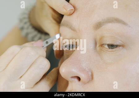 Die Hände des Stylistin zupfen die Augenbrauen mit einer Pinzette auf das Gesicht einer Frau. Nahaufnahme. Gesichtspflege. Stockfoto