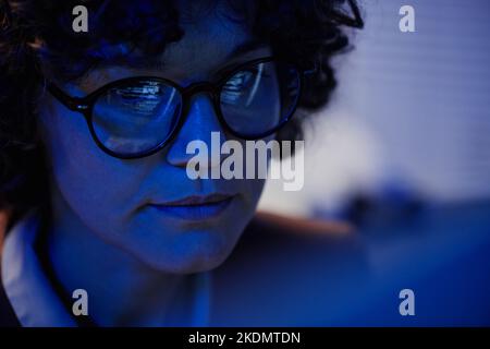 Selektiver Fokus auf das Gesicht einer ernsthaften Geschäftsfrau in Brillen, die spät in der Nacht im Büro vor dem Computer sitzt und Überstunden arbeitet Stockfoto