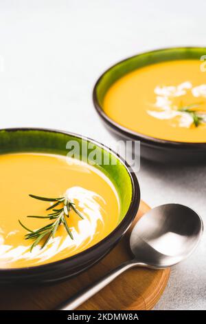 Zwei grüne Schalen mit dickem Butternut-Squash-Bisque, garniert mit schwerer Creme und Rosmarin auf einem grauen Betontisch. Schüssel mit Kürbis und Karotten-Creme so Stockfoto