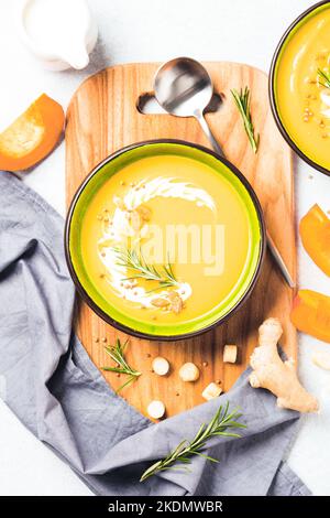 Kürbiscremesuppe garniert mit schwerer Creme und Rosmarin. Dicker Butternut Squash Bisque in grüner Schale auf Holzschneidebrett auf weißem Tisch. Vertic Stockfoto