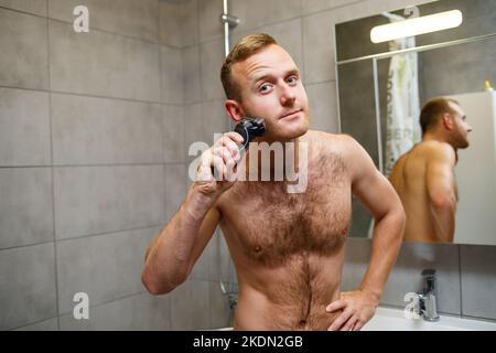 Ein Mann rasiert sein Gesicht mit einem elektrischen Rasierer vor einem Spiegel. Hautirritationen. Badeverfahren Stockfoto