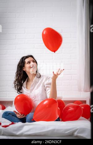 Brunette europäische Mädchen freut sich auf dem Bett mit roten Herz Form Ballons spielen. Morgengeschenk mit Liebesüberraschung am Valentinstag Stockfoto