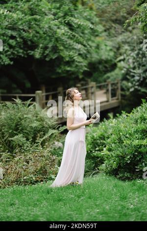 Frau mit rosafarbenem Gown beim Lesen eines Buches in einem idyllischen Garten Stockfoto