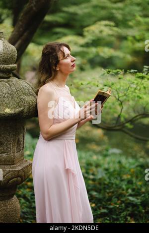 Frau mit rosafarbenem Gown beim Lesen eines Buches in einem idyllischen Garten Stockfoto