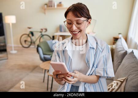 Waist-up-Porträt einer jungen Frau mit Smartphone zu Hause und lächelt fröhlich, gute Nachrichten Stockfoto