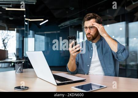 Serious reifen Chef arbeitet im modernen Bürogebäude mit Laptop, Geschäftsmann erhielt schlechte Nachrichten online, Lesen auf dem Smartphone verwirrt wütend und deprimiert Mann bei der Arbeit. Stockfoto